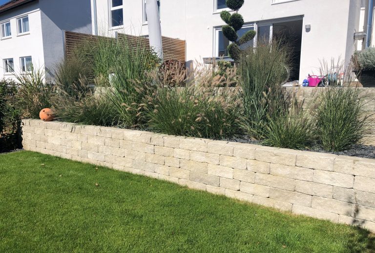 Niedrige helle Gartenmauer mit Gräsern und Stauden oberhalb einer Rasenfläche.