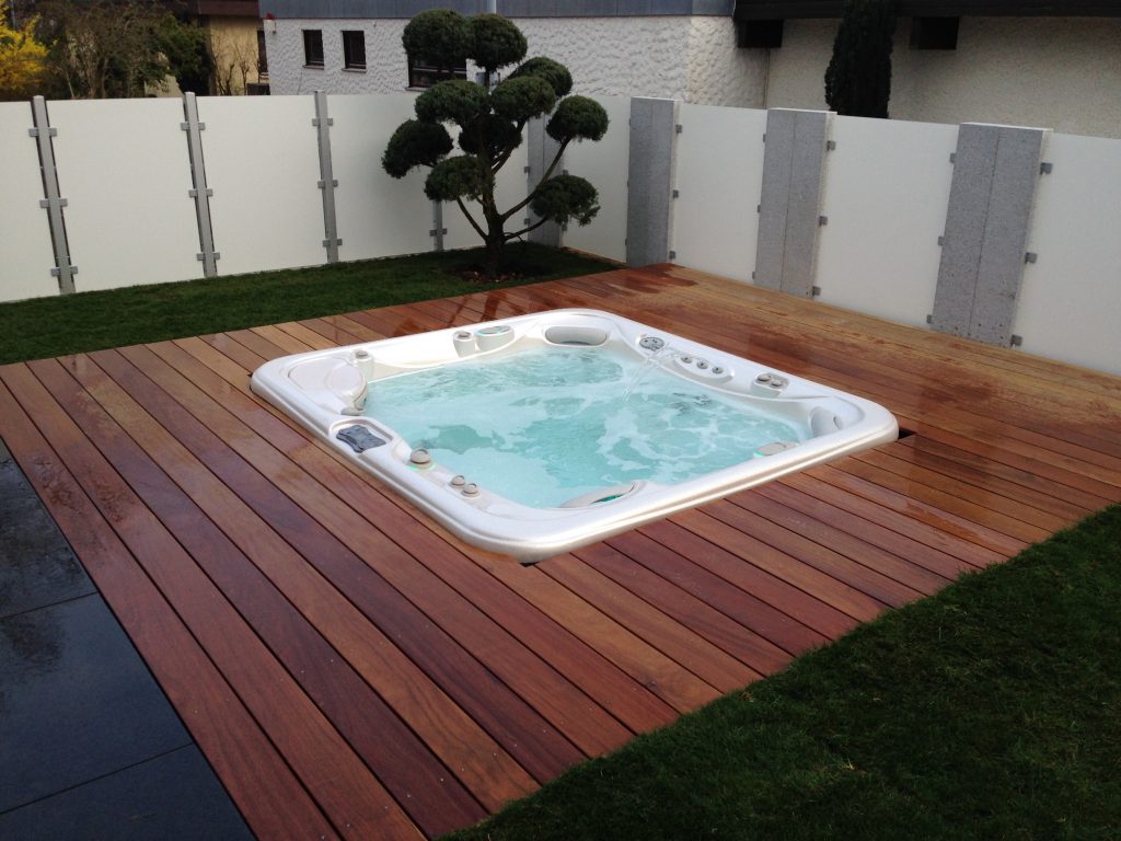 In eine Holzterrasse eingelassener Whirlpool vor hellem Sichtschutz.