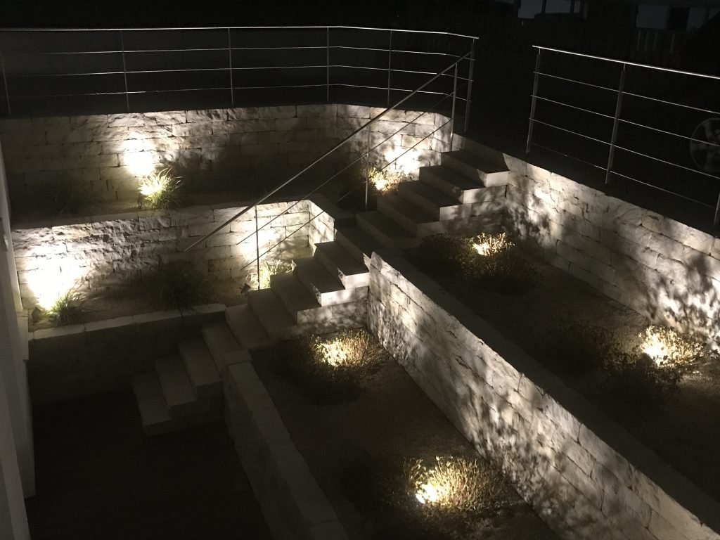 Gestaffelte Natursteinmauern mit punktueller Beleuchtung bei Nacht.