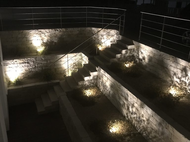 Gestaffelte Natursteinmauern mit punktueller Beleuchtung bei Nacht.