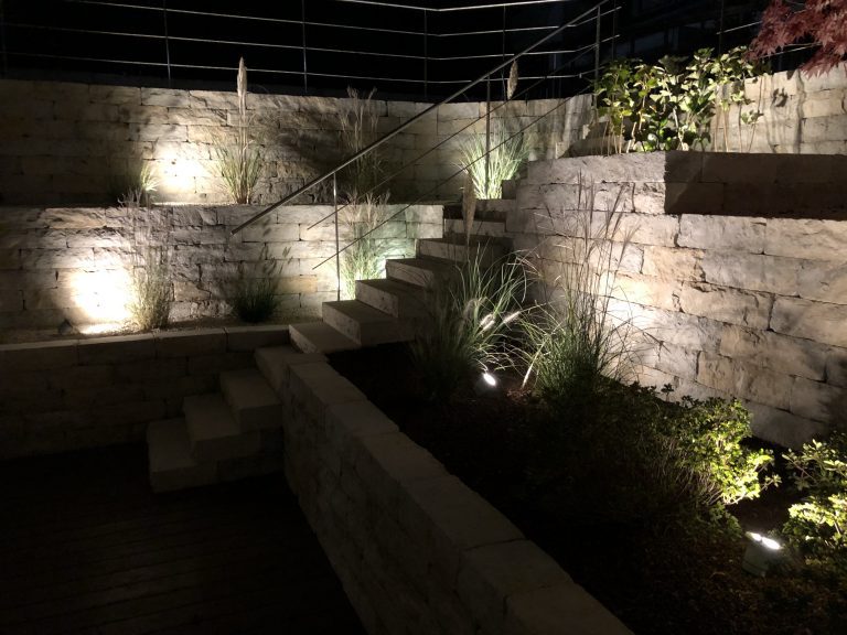 Beleuchteter Lichthof mit bepflanzten Natursteinmauern und Treppenstufen.