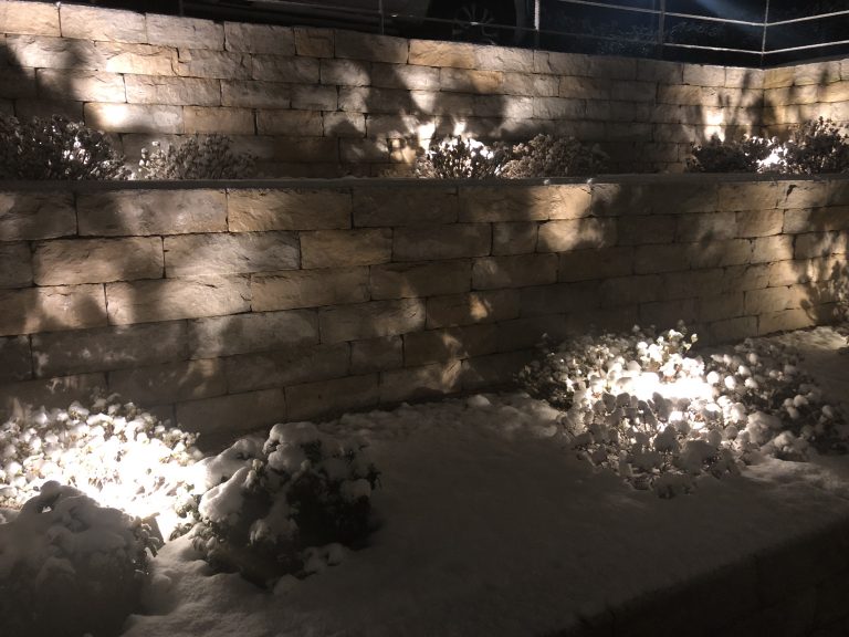 Natursteinmauer und verschneites Gartenbeet mit warmen Lichtakzenten.