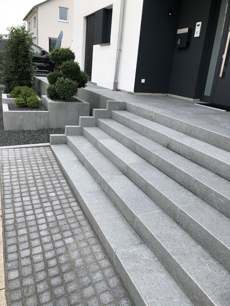 Breite Eingangstreppe mit dunklem Natursteinpflaster und bepflanzten Randbereichen.