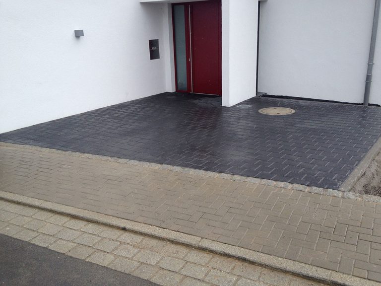 Dunkel gepflasterter Hauseingang mit hellem Randabschluss.