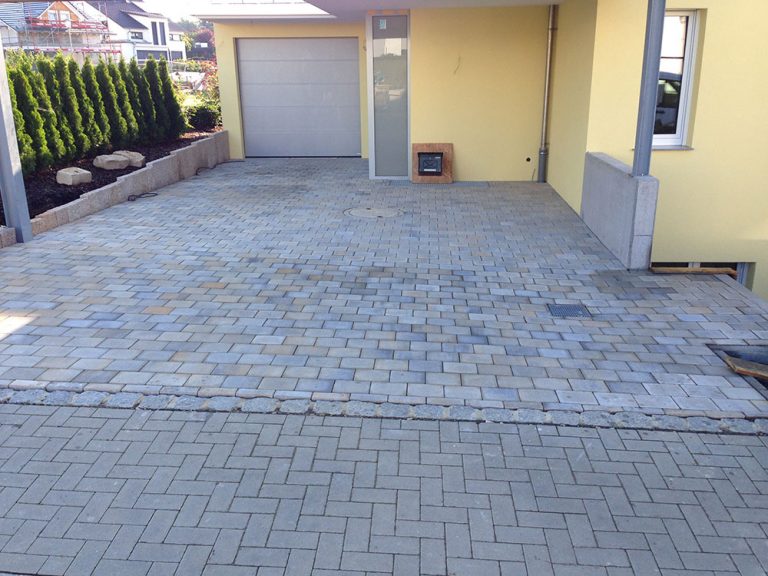 Grau gepflasterte Einfahrt vor einer Garage an einem gelben Wohnhaus.