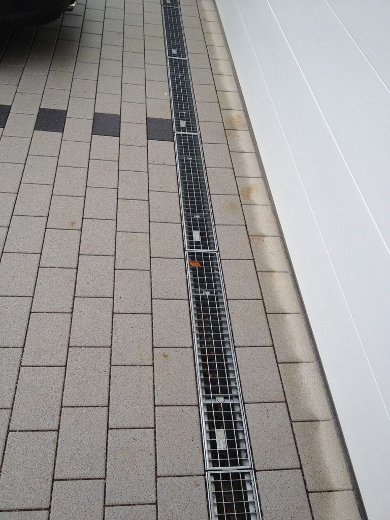 Pflasterbelag mit gerader Entwässerungsrinne entlang einer Hauswand.