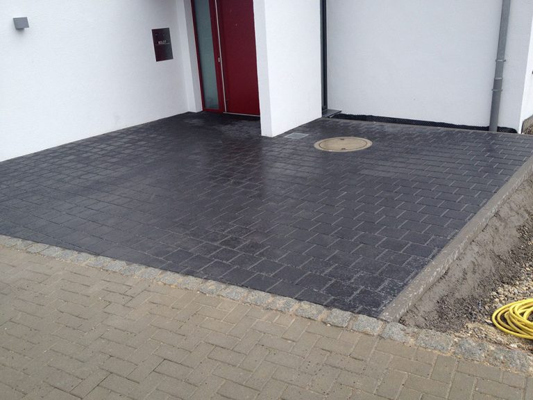 Dunkles Pflaster vor einer Haustür mit passgenau eingefasstem Schachtdeckel.