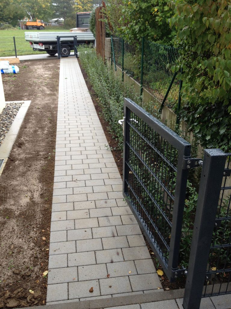 Schmaler heller Pflasterweg entlang eines Metallzauns.