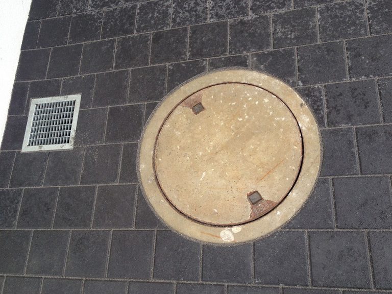 Schachtdeckel und kleiner Ablauf in anthrazitfarbenem Pflaster.