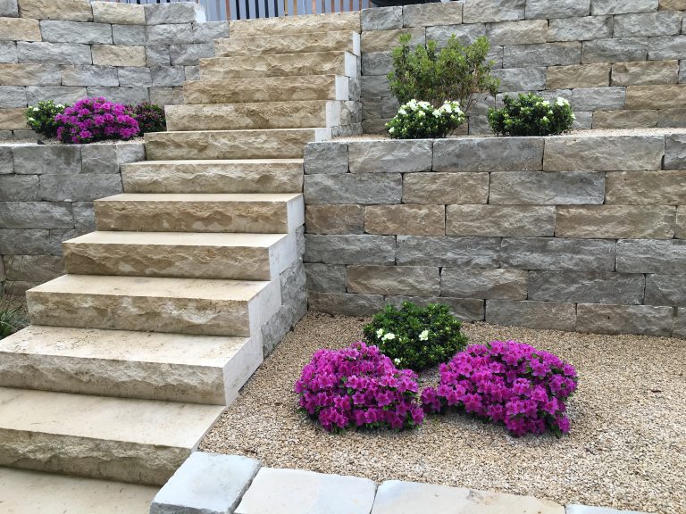 Natursteintreppe zwischen bepflanzten Mauern mit violetten Blüten.