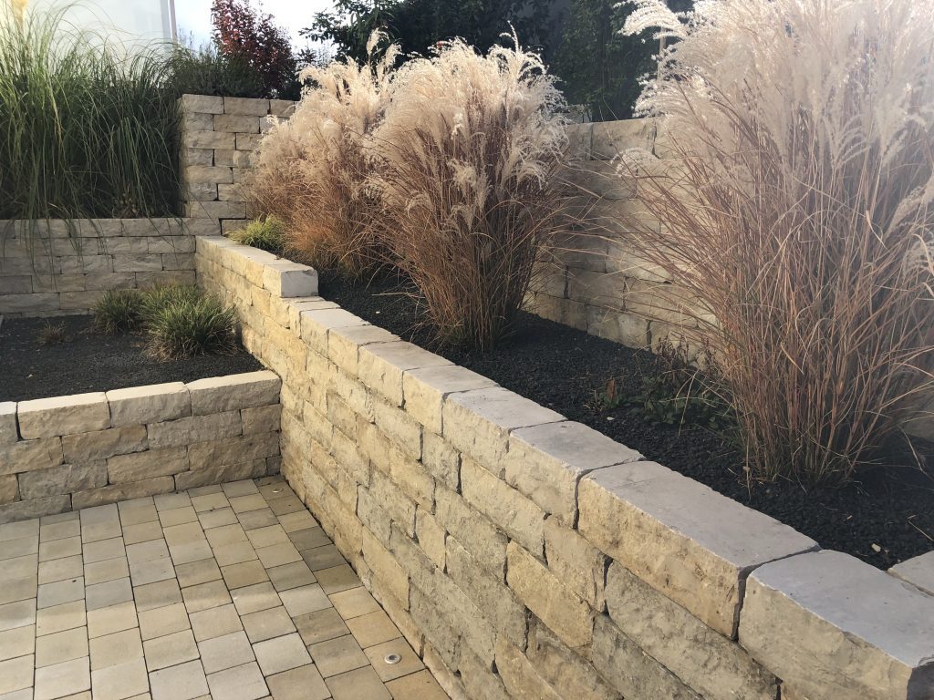 Natursteinmauer mit üppigen Ziergräsern oberhalb einer gepflasterten Fläche.