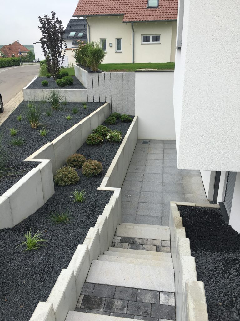 Außentreppe und schmaler Hauszugang zwischen grauen Einfassungen und bepflanzten Beeten.