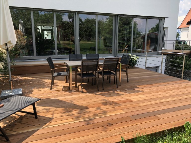 Holzterrasse mit Esstisch, Stühlen und Liege vor großen Fenstern.