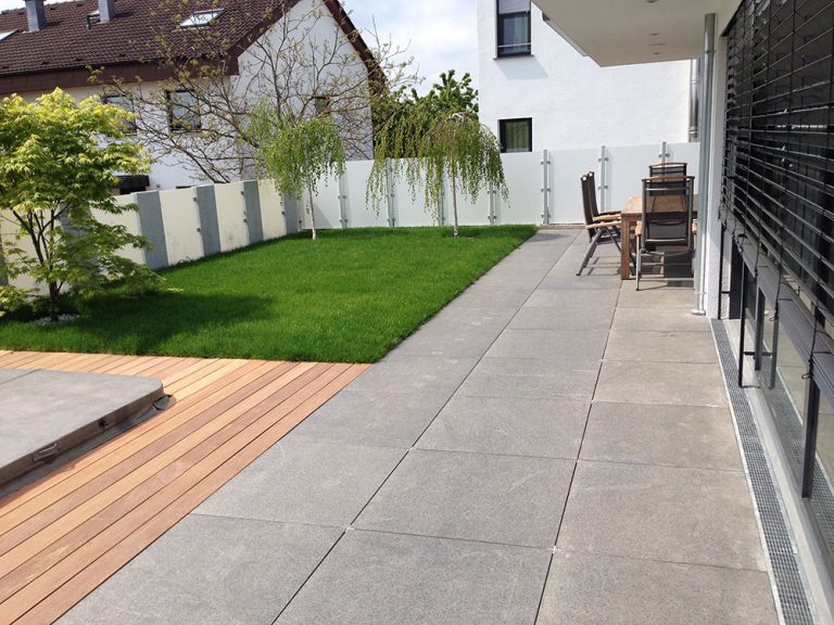 Helle Terrasse entlang eines Wohnhauses mit Rasen und Holzdeck.