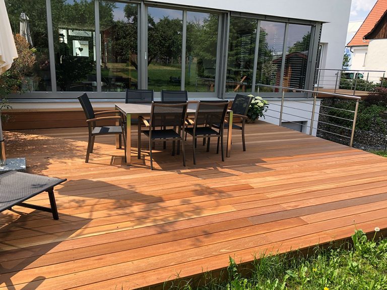 Warme Holzdielen-Terrasse mit Essgruppe und Liege an einer Glasfront.