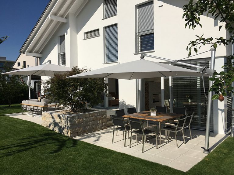 Terrasse mit Sonnenschirmen und Gartenmöbeln zwischen Wohnhaus und Rasenfläche.