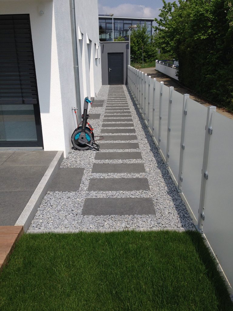 Gerader Trittplattenweg im Kies entlang einer Hausfassade und eines Sichtschutzes.