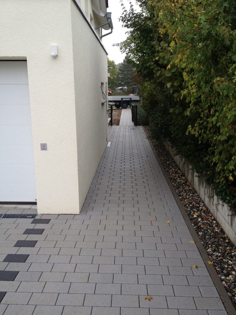 Gerader Pflasterweg zwischen Hauswand und dichter Hecke.