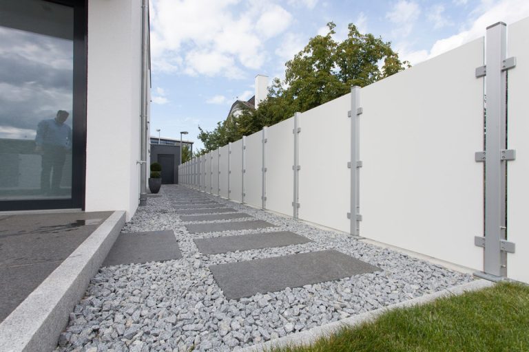 Schmaler Kies- und Plattenweg entlang eines hellen Glas-Sichtschutzes.