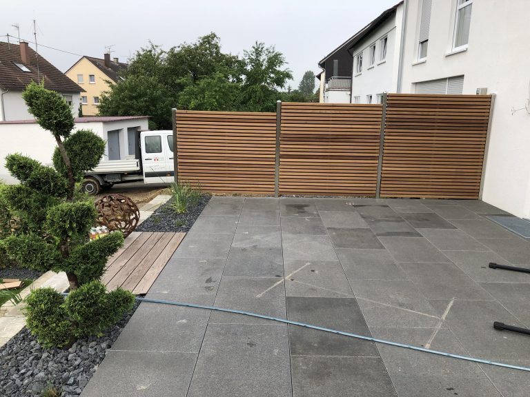 Terrassenplatten mit Holzlamellen-Sichtschutz und geschnittenen Ziergehölzen.