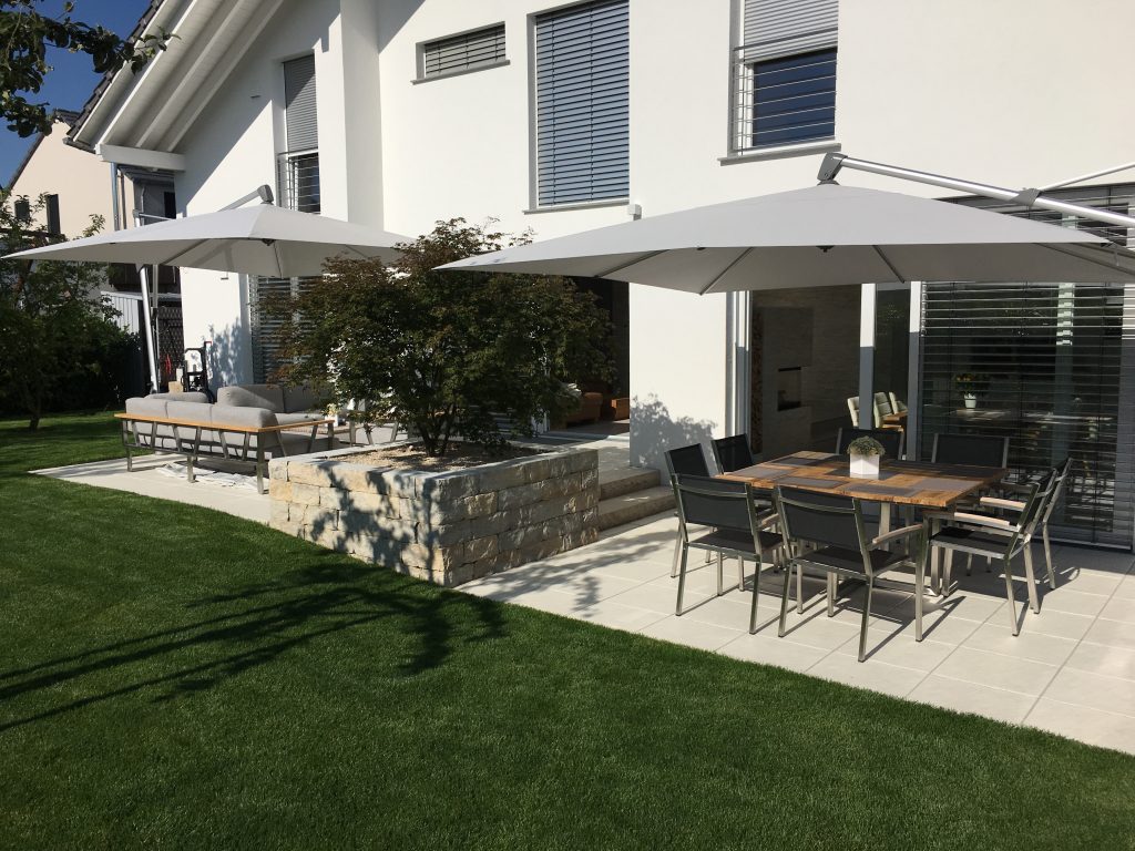 Terrasse mit großen Sonnenschirmen, Sitzgruppen und angrenzender Rasenfläche vor einem Wohnhaus.