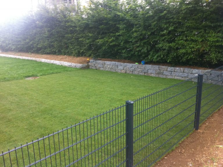 Rasenfläche mit Metallzaun und niedriger Natursteinmauer vor einer Hecke.