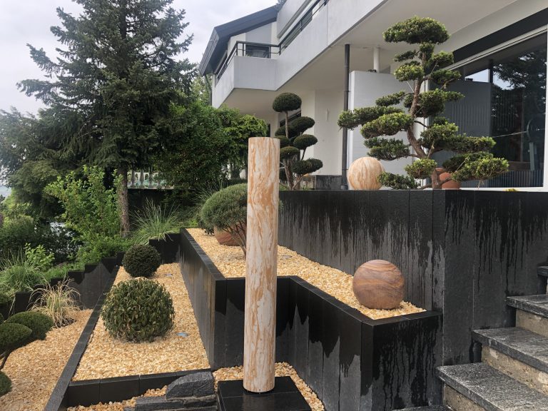 Vorgarten mit dunklen Pflanztrögen, Ziergehölzen und hellem Kies.