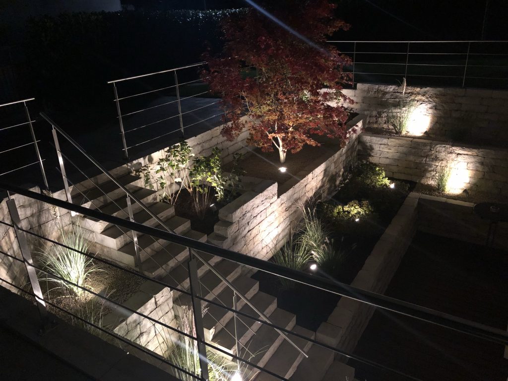 Beleuchteter Lichthof mit Natursteinmauern, Treppe und rotem Fächerahorn.