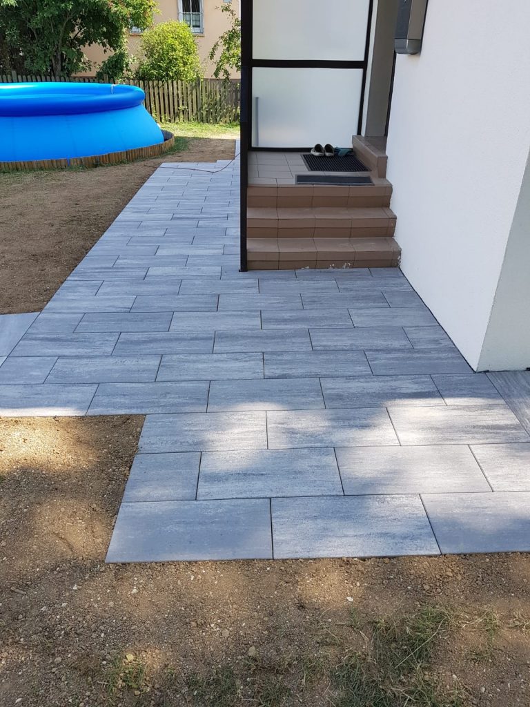 Großformatiger Plattenbelag rund um eine kleine Holztreppe am Haus.