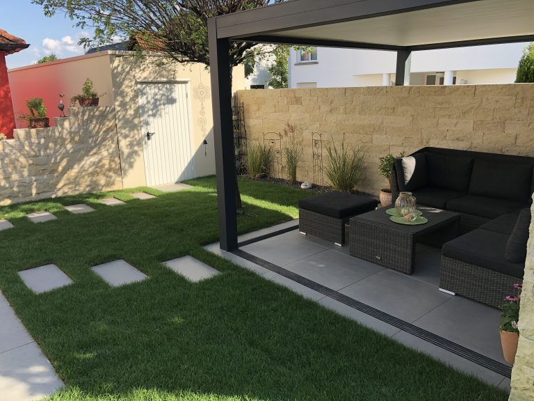 Überdachter Gartensitzplatz mit dunkler Lounge, Naturstein-Sichtschutz und Trittplatten im Rasen.