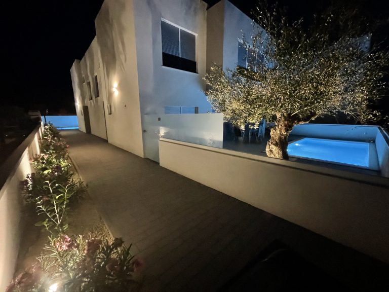 Abendlicher Hausweg mit beleuchteten Beeten und blau leuchtendem Pool.