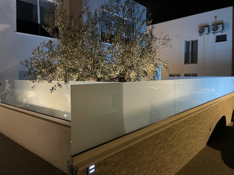 Abendlicher Terrassenbereich mit beleuchtetem Olivenbaum hinter hellen Glaselementen.