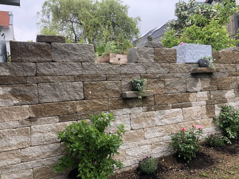 Natursteinmauer mit kleinen Pflanznischen und bepflanztem Beet davor.