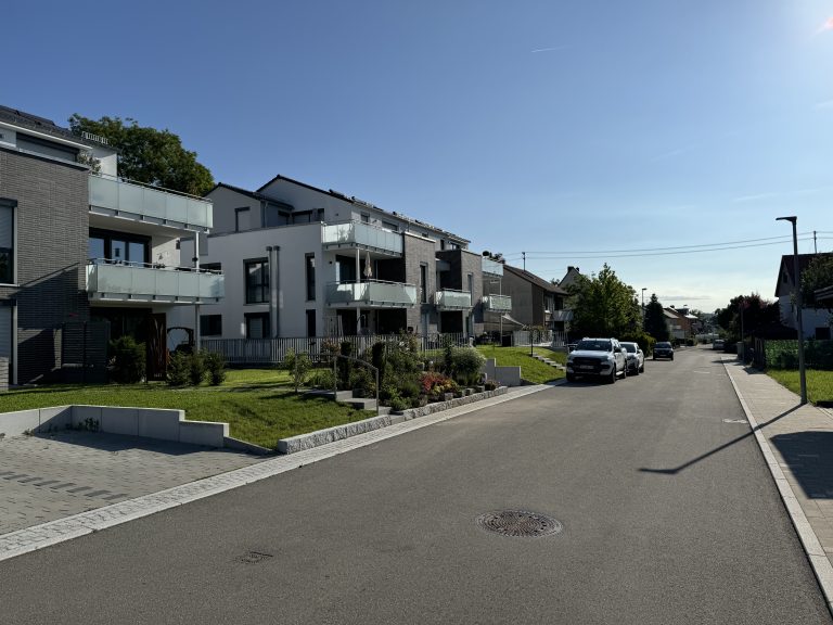 Straßenansicht einer Wohnanlage mit begrünten Vorgärten und Zugangswegen.
