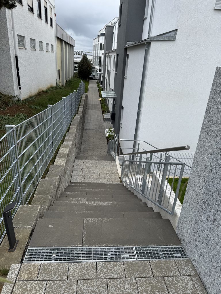Außentreppe mit Metallgeländer und befestigtem Weg entlang einer Hauswand.