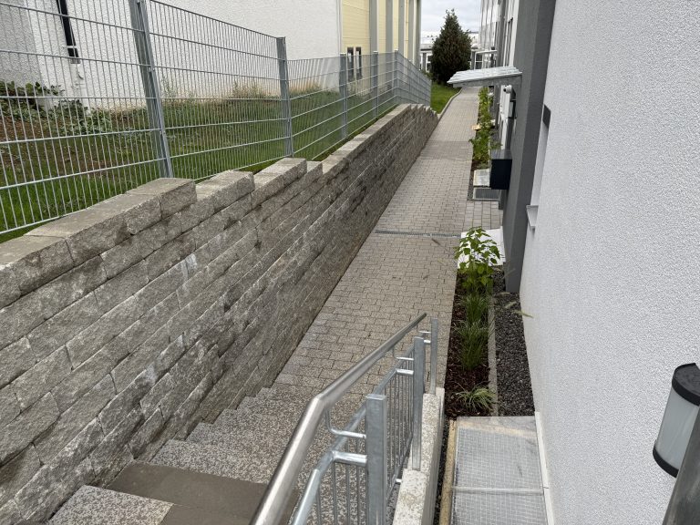 Naturstein-Stützmauer neben einem abgesenkten Hauszugang mit Geländer.