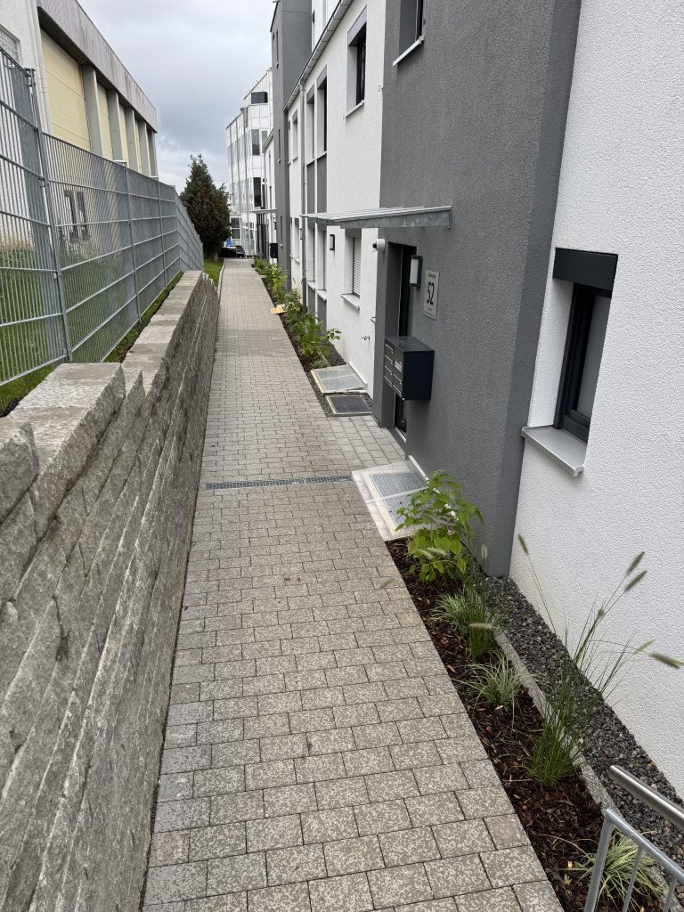 Schmaler gepflasterter Hausweg entlang einer hohen Naturstein-Stützmauer.