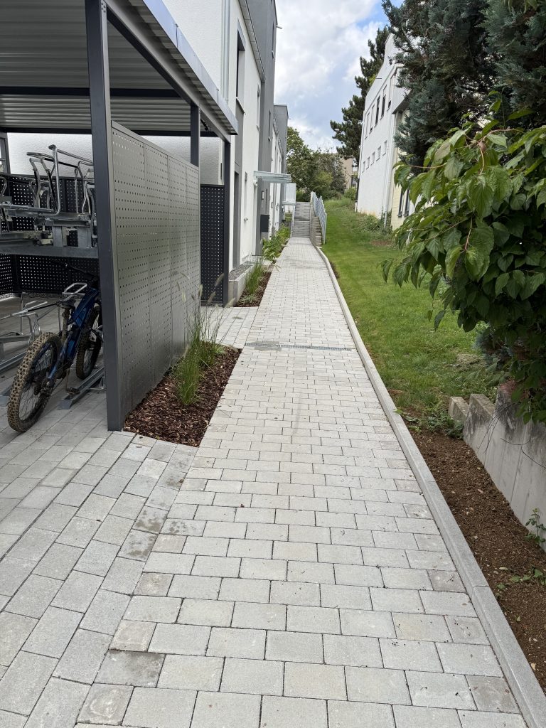 Heller Pflasterweg zwischen Hauswand und einer schmalen Pflanzfläche.