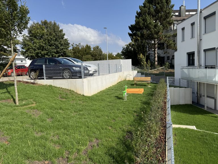 Rasenböschung und Stützmauer zwischen Straße und Wohngebäude.