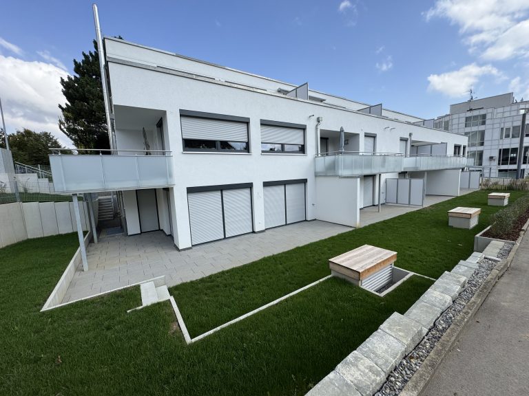Außenanlage eines modernen Wohngebäudes mit Rasen, Plattenwegen und niedrigen Einfassungen.