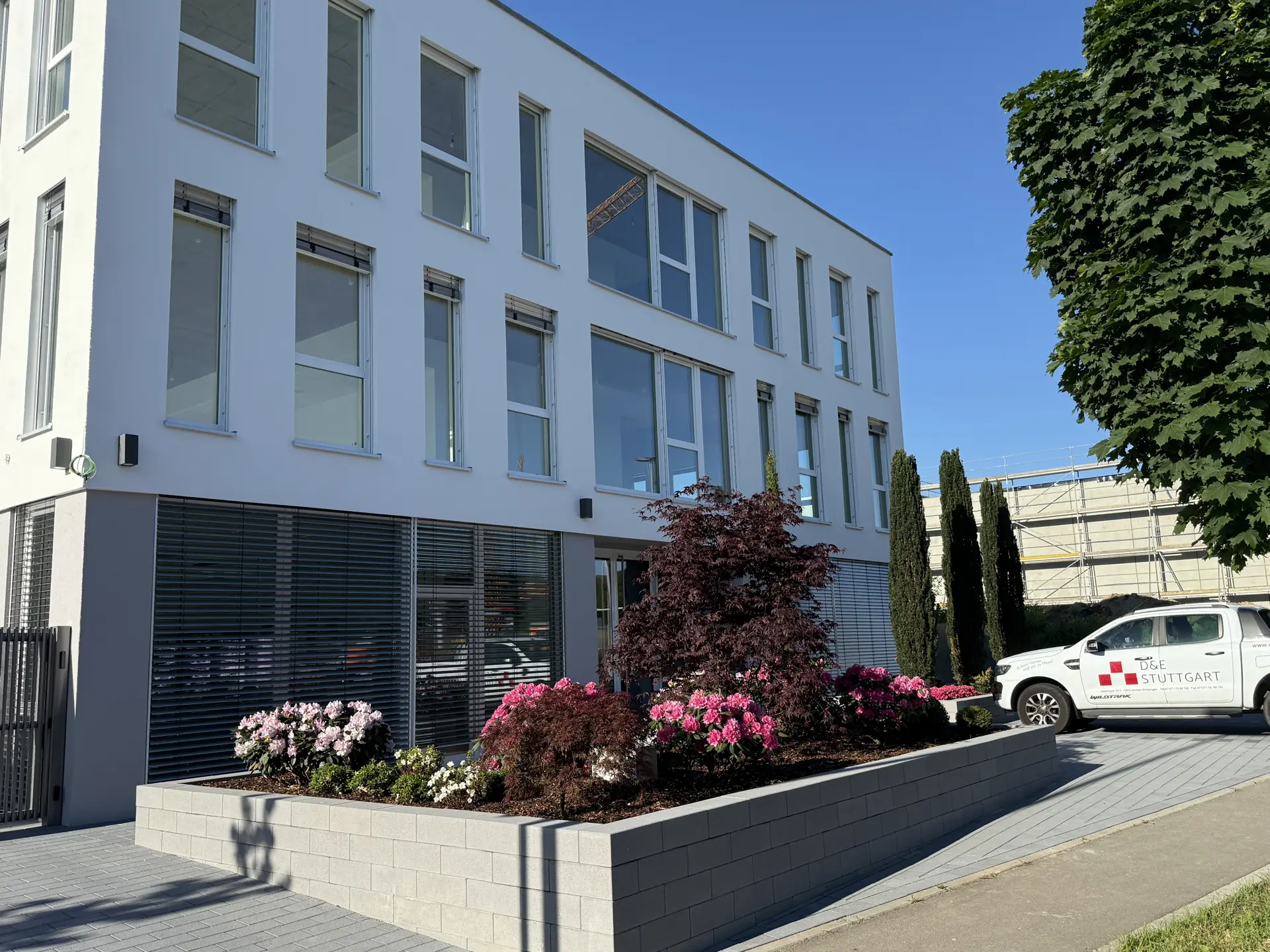 Gesamtansicht einer bepflanzten Außenanlage mit grauer Pflanzmauer und dem Firmenfahrzeug von D&E Stuttgart.