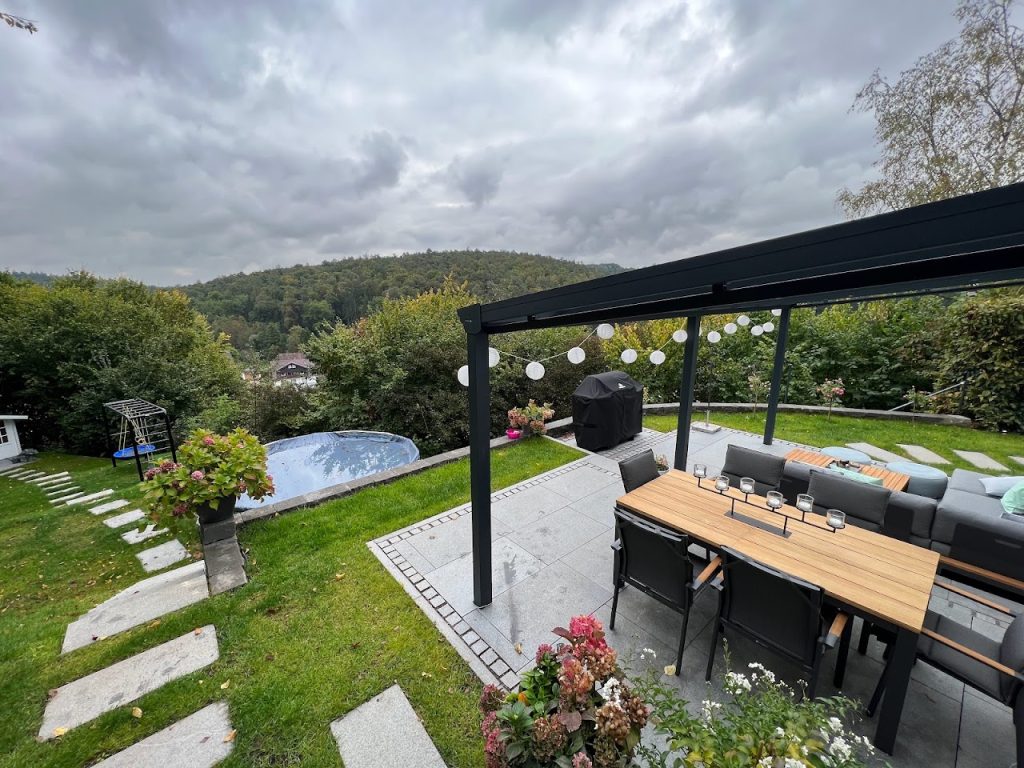 Essplatz unter einer dunklen Pergola mit hellen Terrassenplatten, Rasen und Trittplatten im Garten.