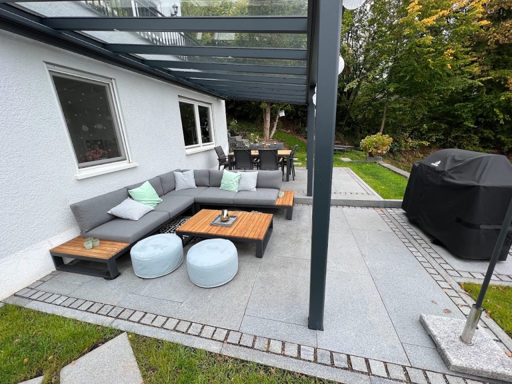 Graue Lounge unter einer dunklen Pergola auf einer hell gepflasterten Terrasse mit Essplatz und Garten.